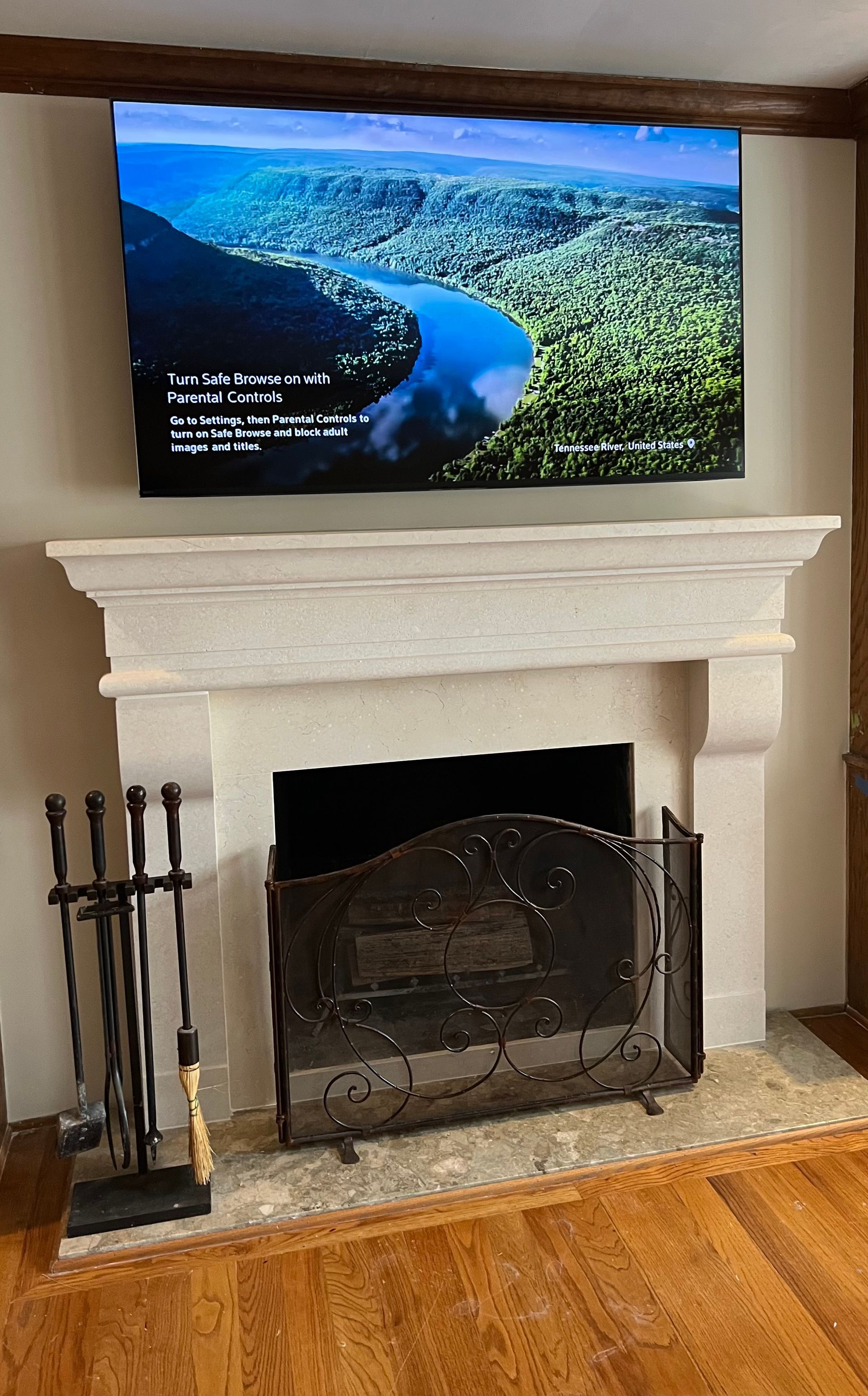 TV above a fireplace displaying a scenic river view. Black fire screen and tools are in front of the fireplace.