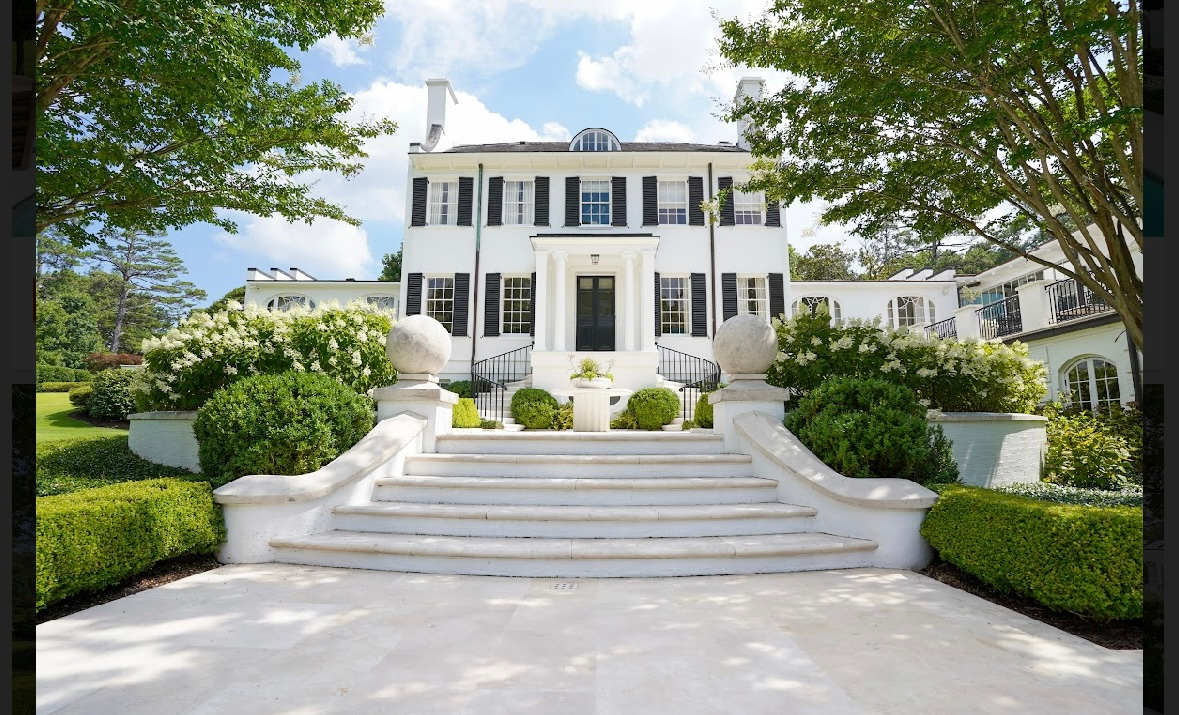 White mansion with steps, green hedges, and blue sky.