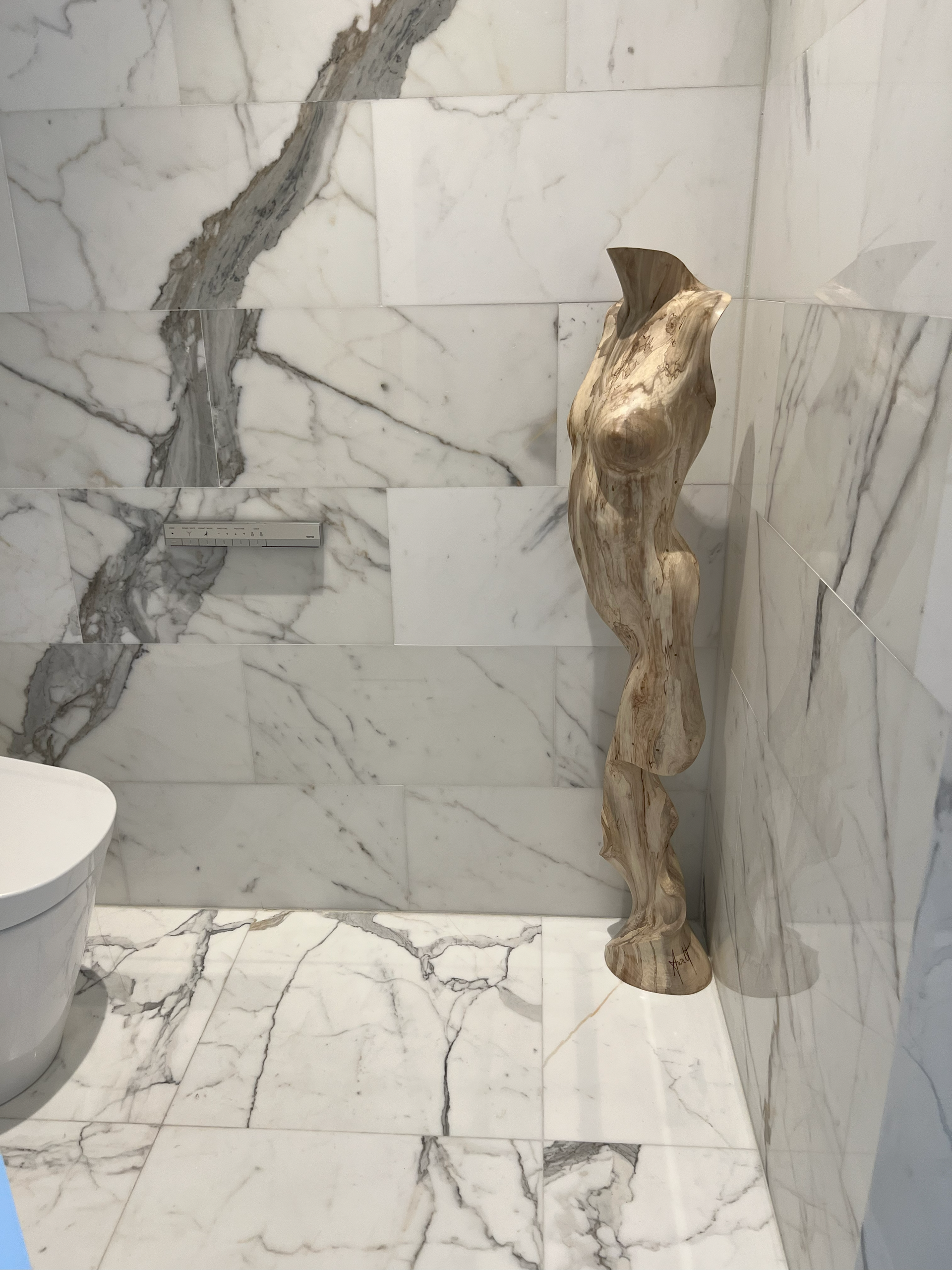 Statue of a torso in a marble-tiled bathroom. White and gray marble wall with a toilet.