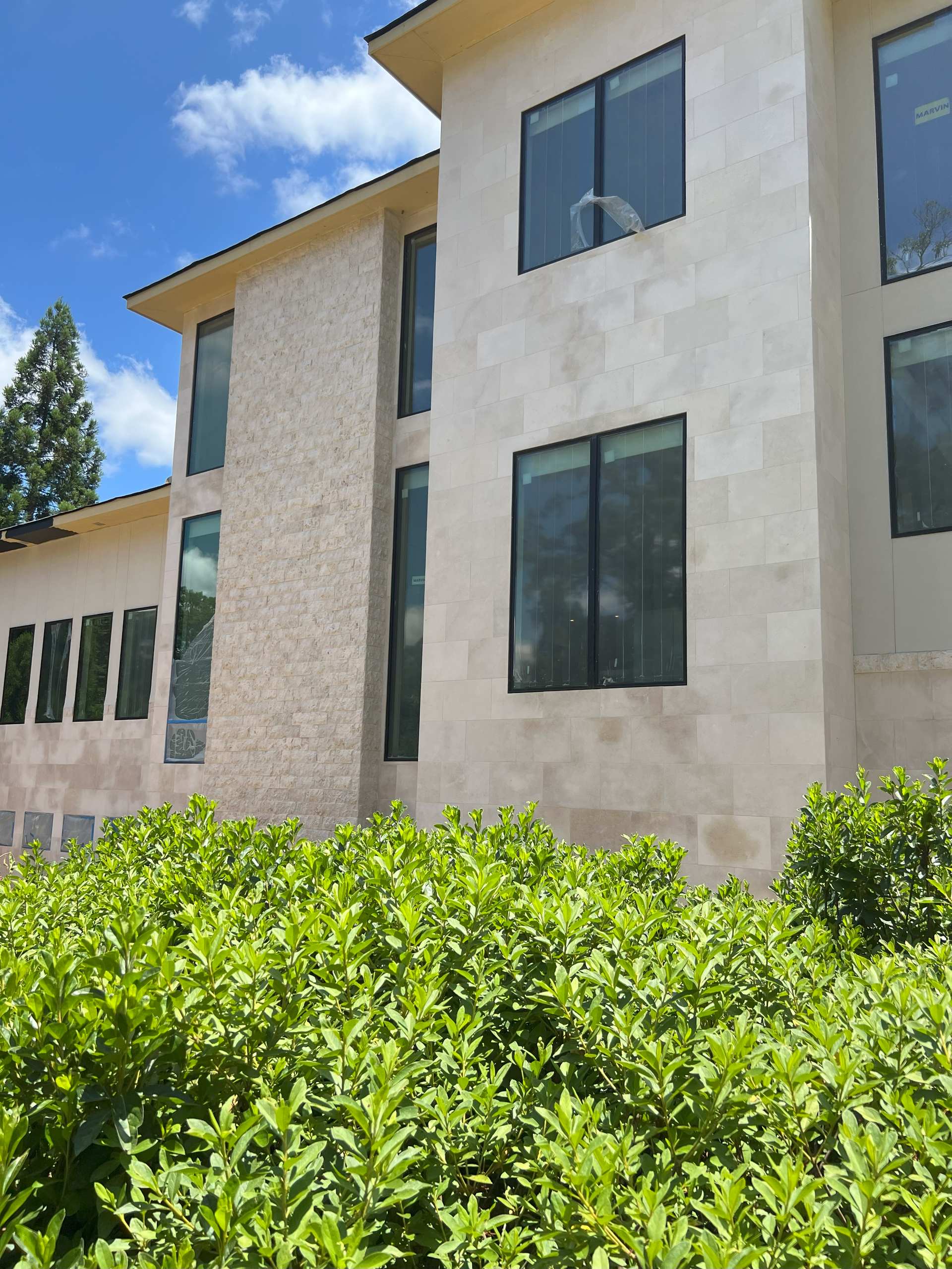 Beige building with dark windows behind green shrubs under a blue sky.