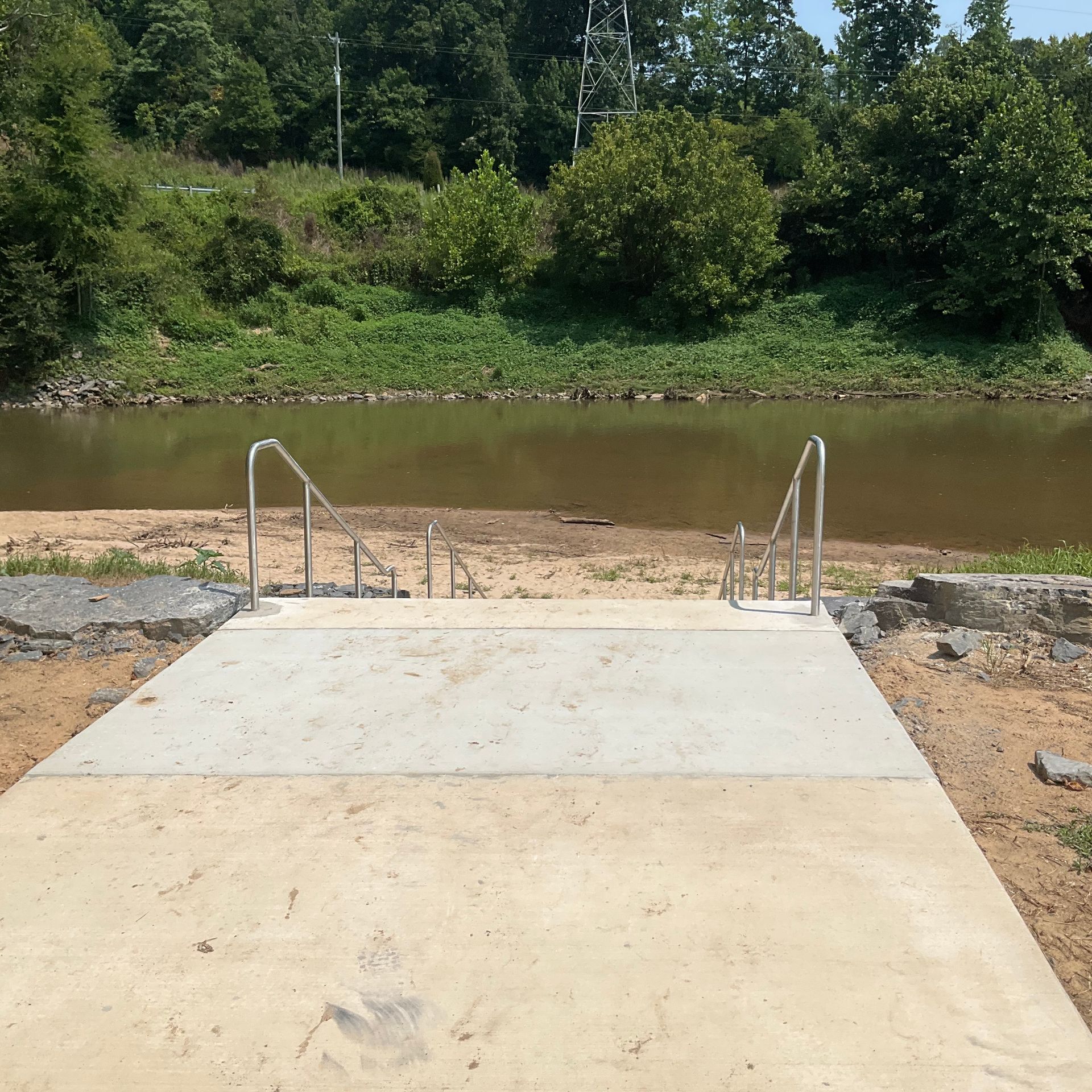Concrete ramp leading to a muddy riverbank with handrails, backed by green trees and shrubs.