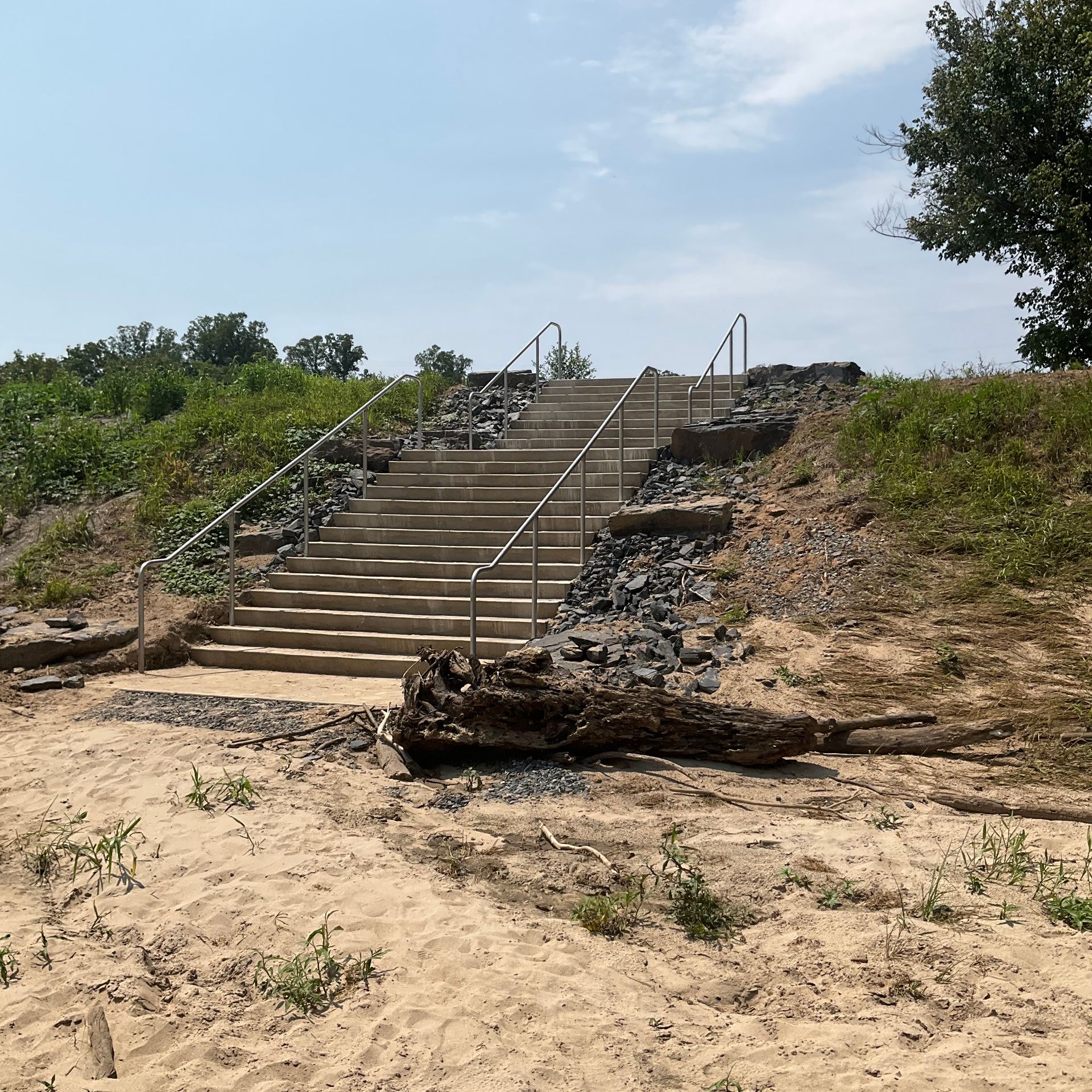 Concrete steps up a sandy hillside with metal handrails, rocky sides, and a fallen log in the foreground