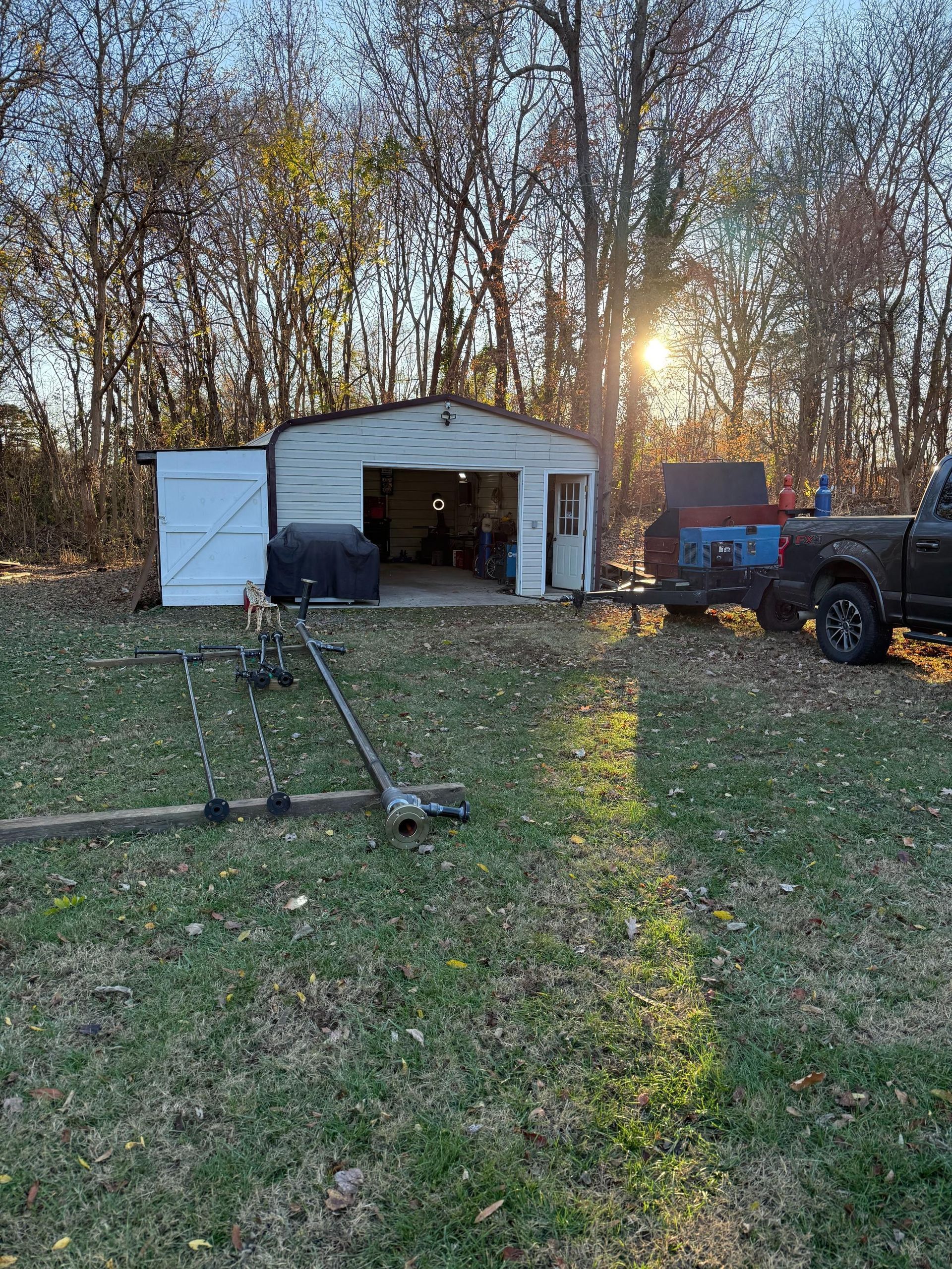 Shed with open doors, a vehicle, and a truck parked outside in a grassy area with trees and sunlight.