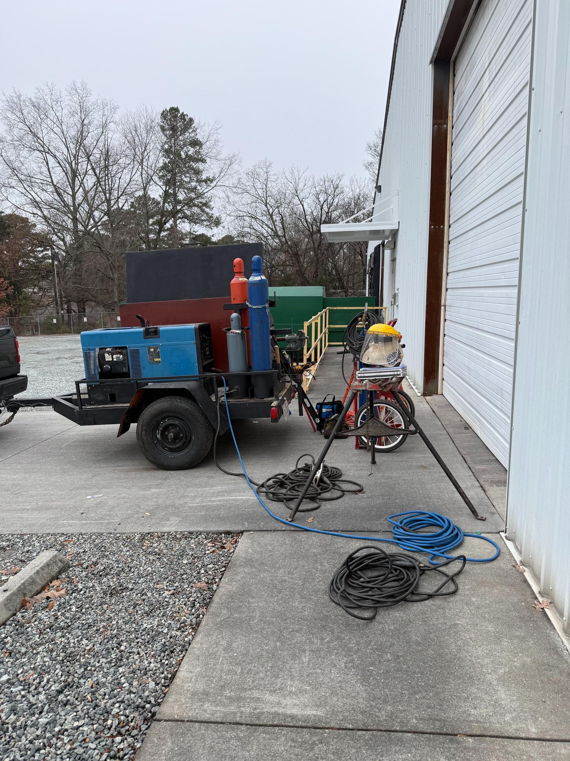 Welding trailer parked near a building. Includes welding equipment, oxygen tanks, and a bicycle. Grey sky.