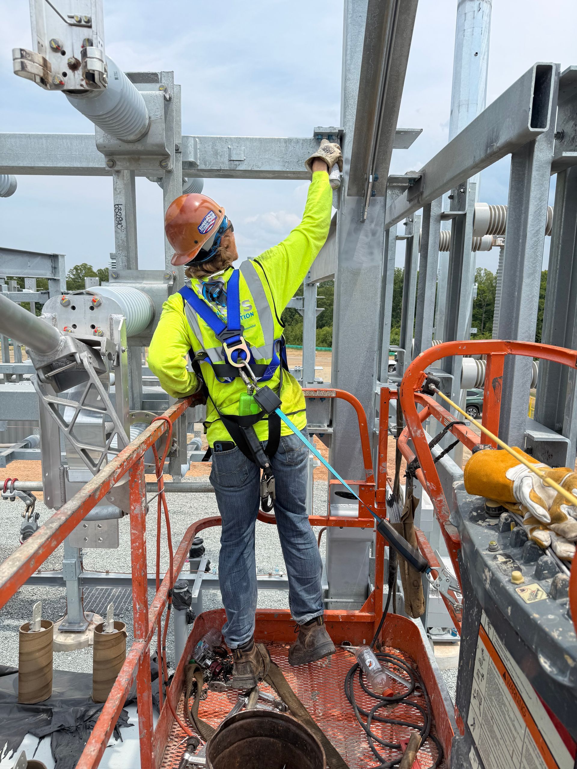 Worker in neon safety gear standing on an industrial lift platform, reaching up in a refinery.