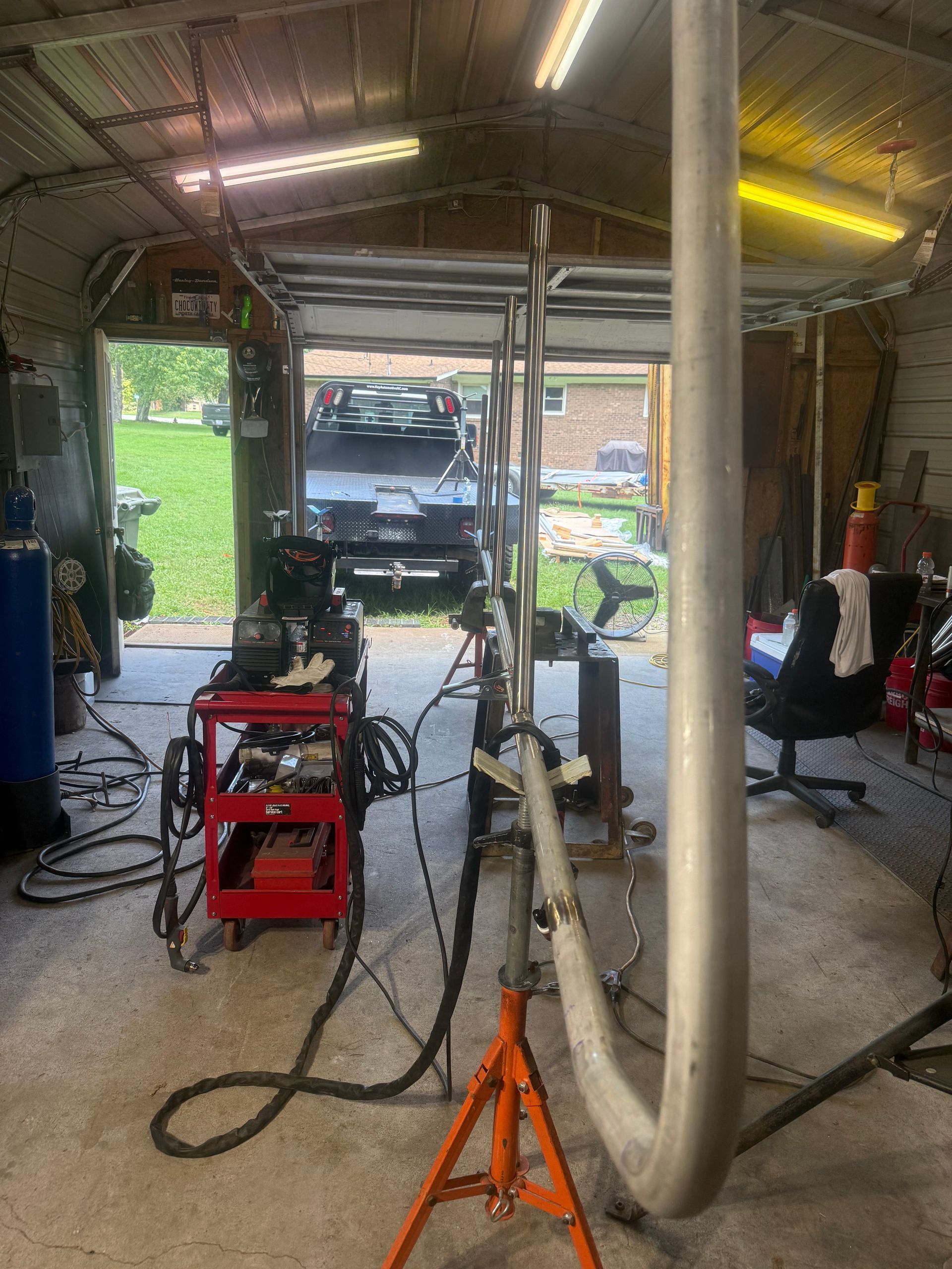 Garage workspace with welding equipment, hoses, and an orange jack stand near an open door