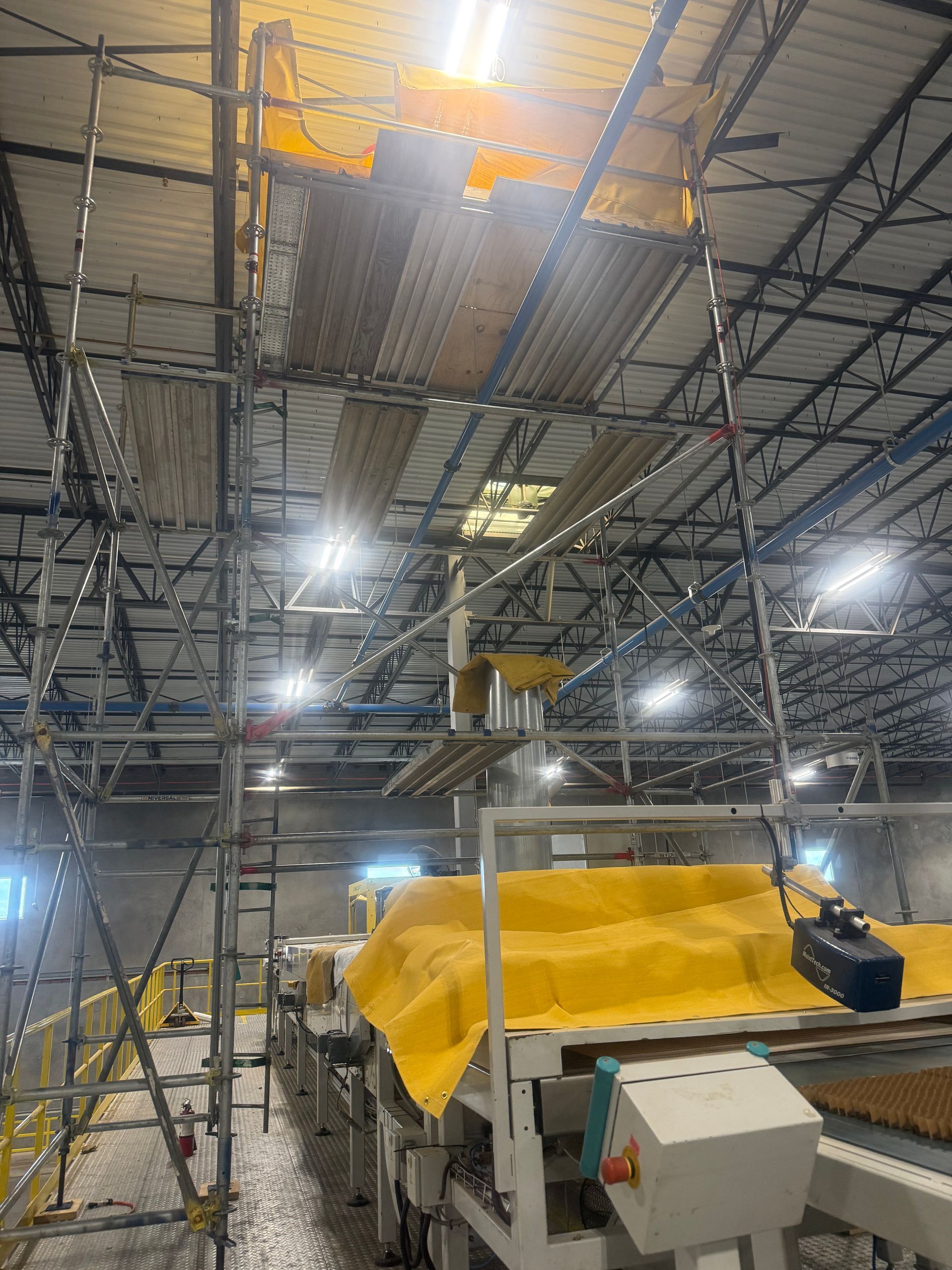 Scaffolding and industrial equipment inside a warehouse, with yellow-covered machinery under bright lights.