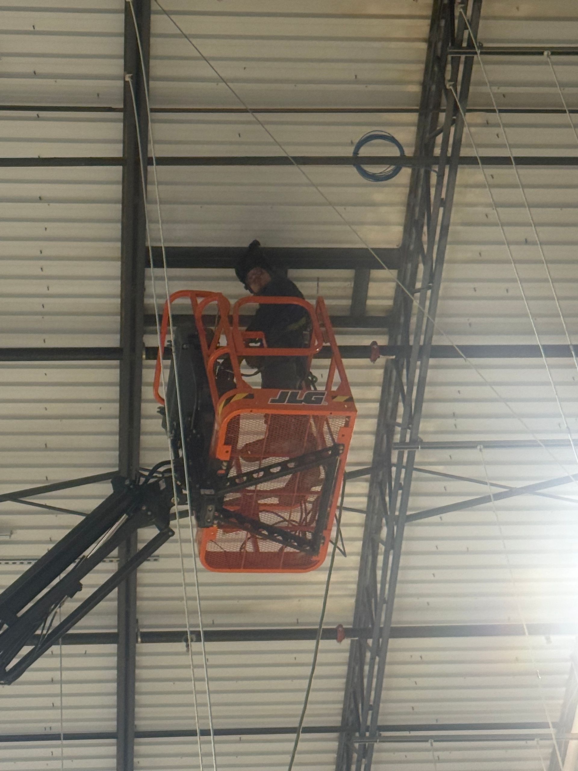 Worker in an orange lift basket inspecting a ceiling inside a warehouse.