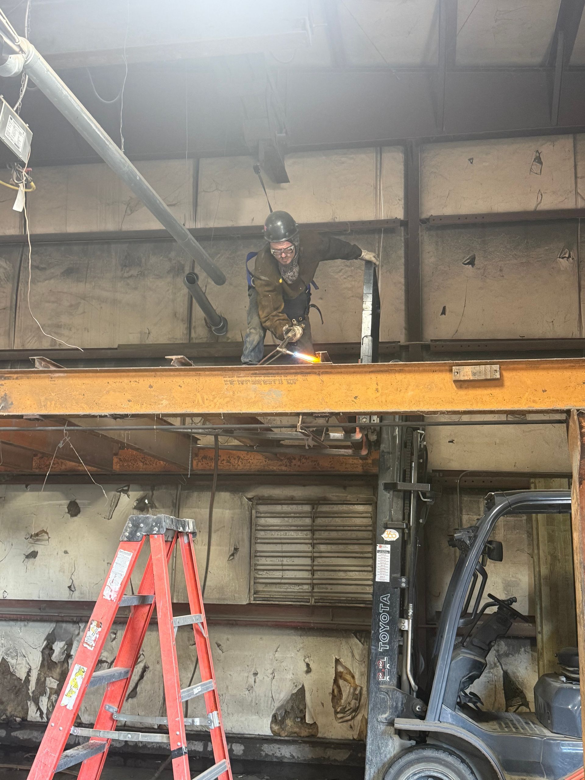 Worker on an orange platform under a large industrial structure, with a red ladder and machinery below.