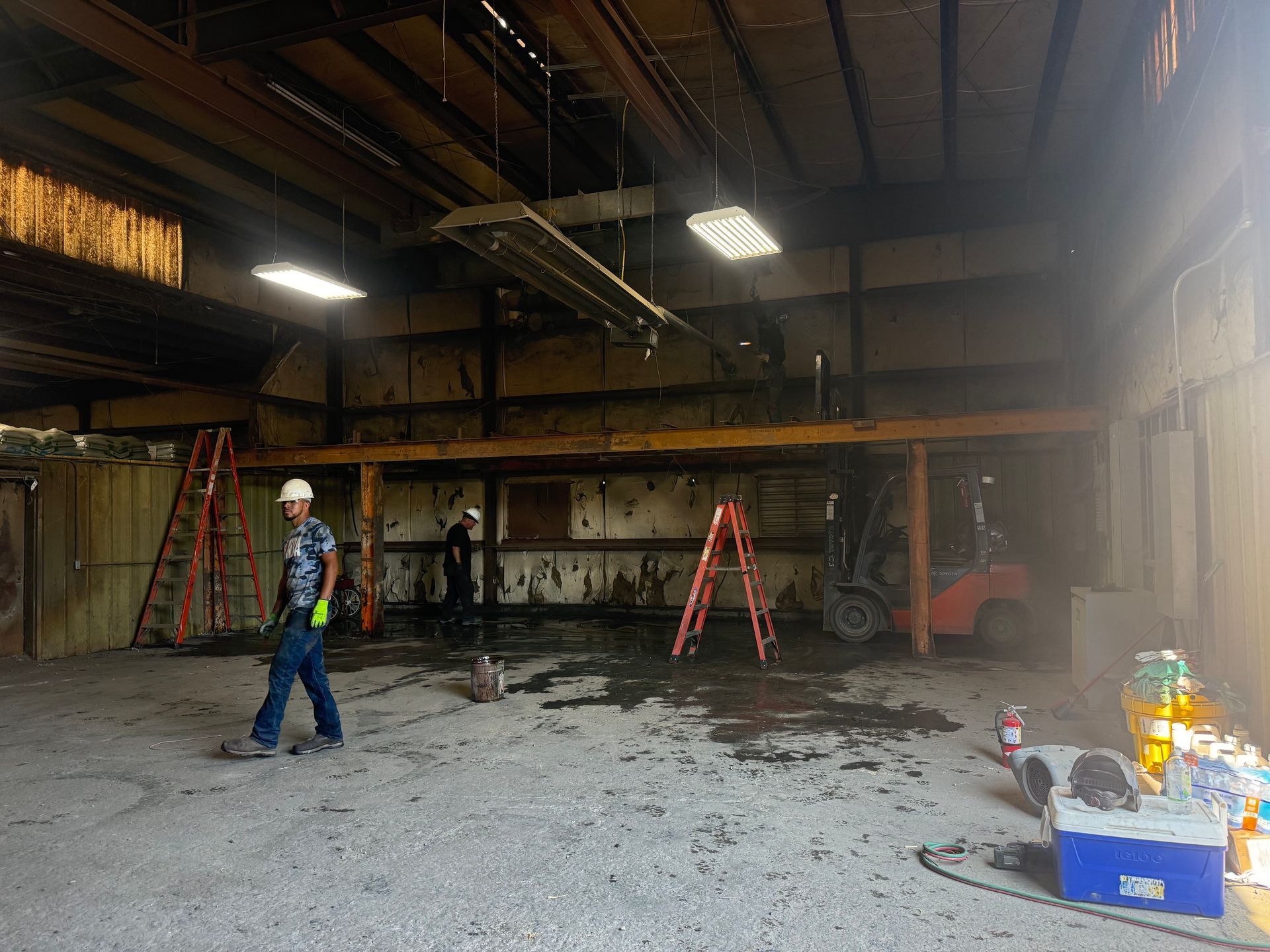 Worker in a large industrial warehouse with ladders, debris, and scattered equipment on the floor.