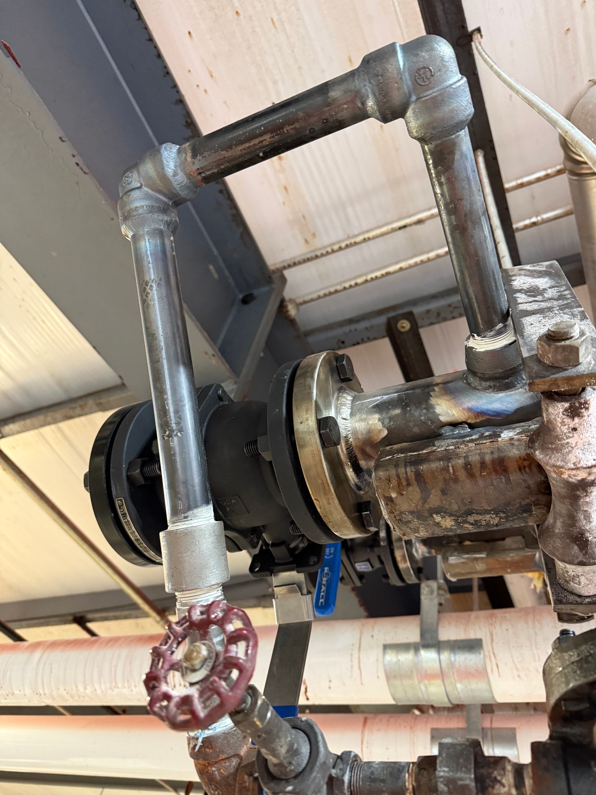 Industrial piping system with valves and a black component, possibly a pump, in an indoor setting.