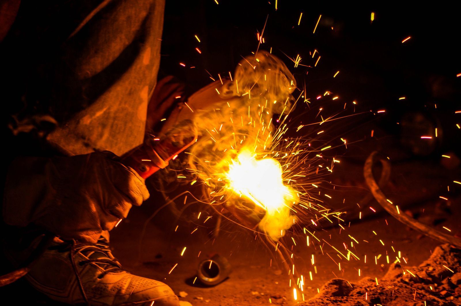 Welder wearing gloves using a welding torch on metal pipes. Sparks and bright light are present.