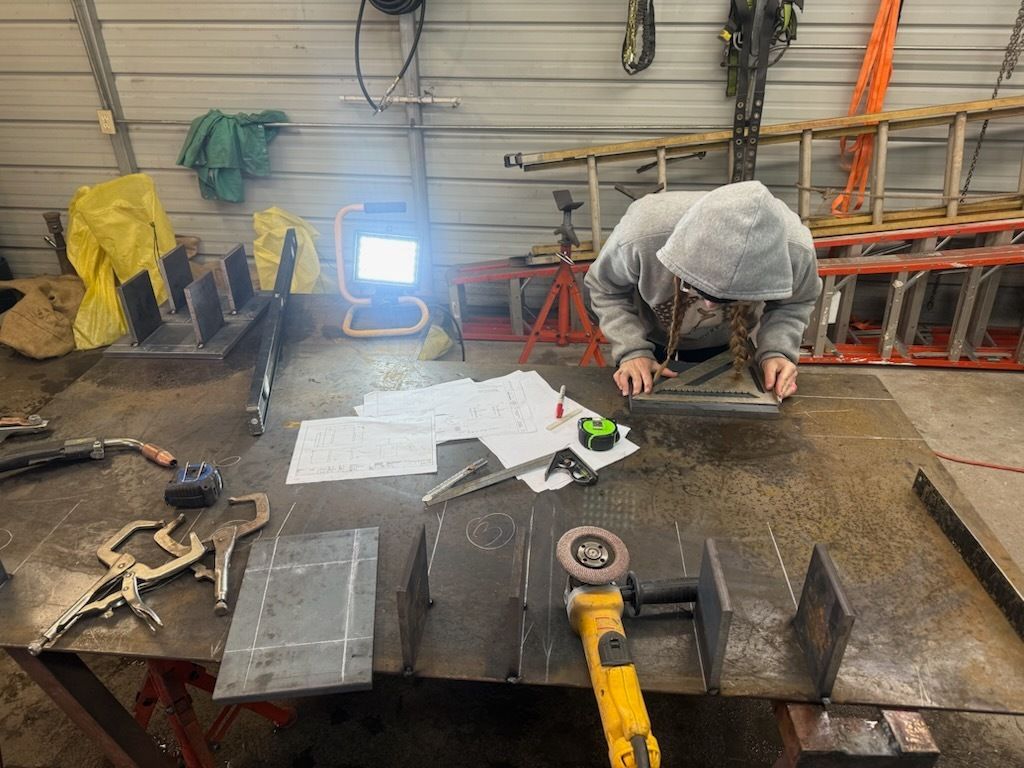 Person in hooded sweatshirt working with metal, using a square and measuring tools on a workbench in a workshop setting.