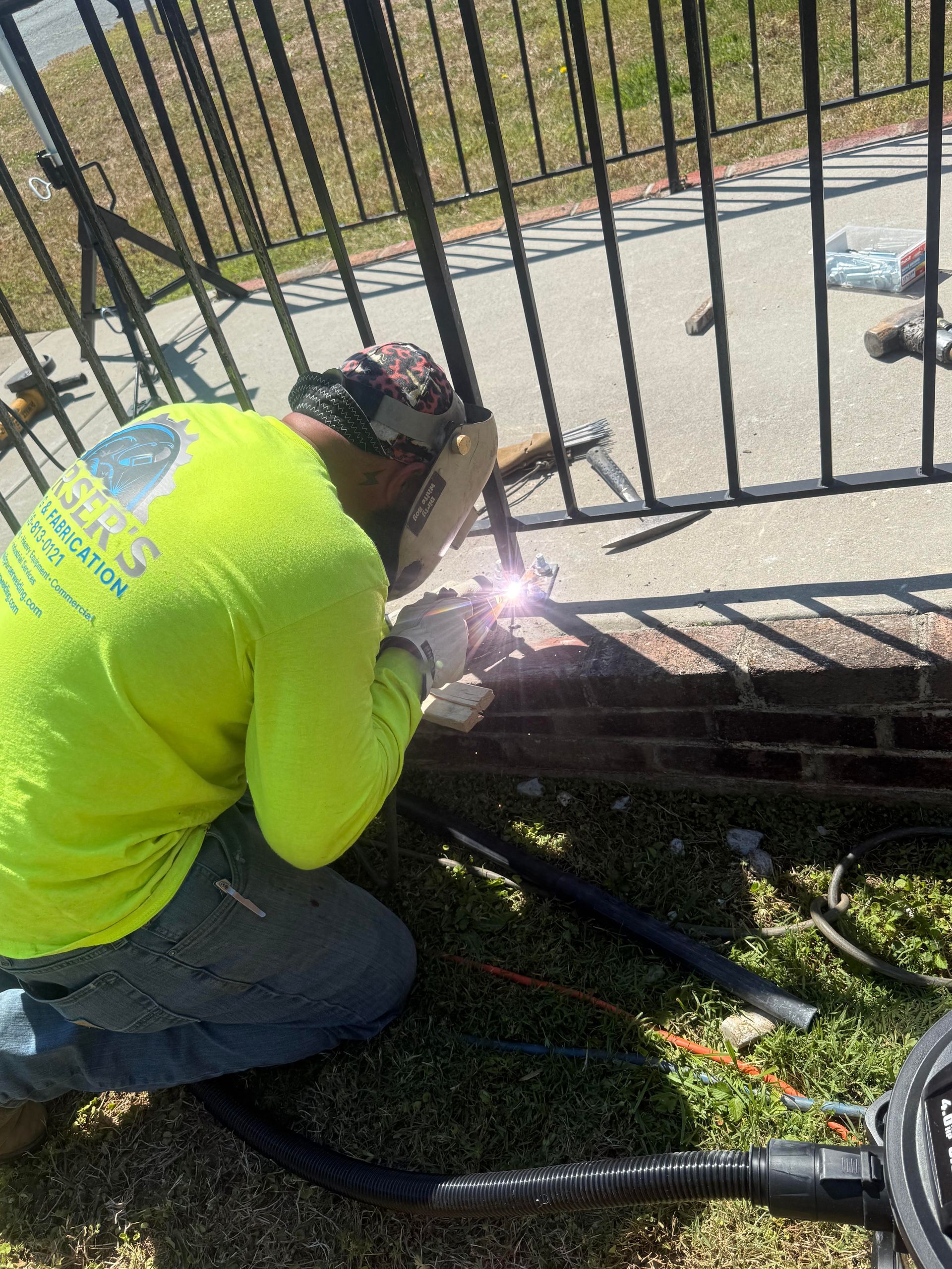 Worker in neon shirt welding a black metal fence, with sparks flying outdoors.