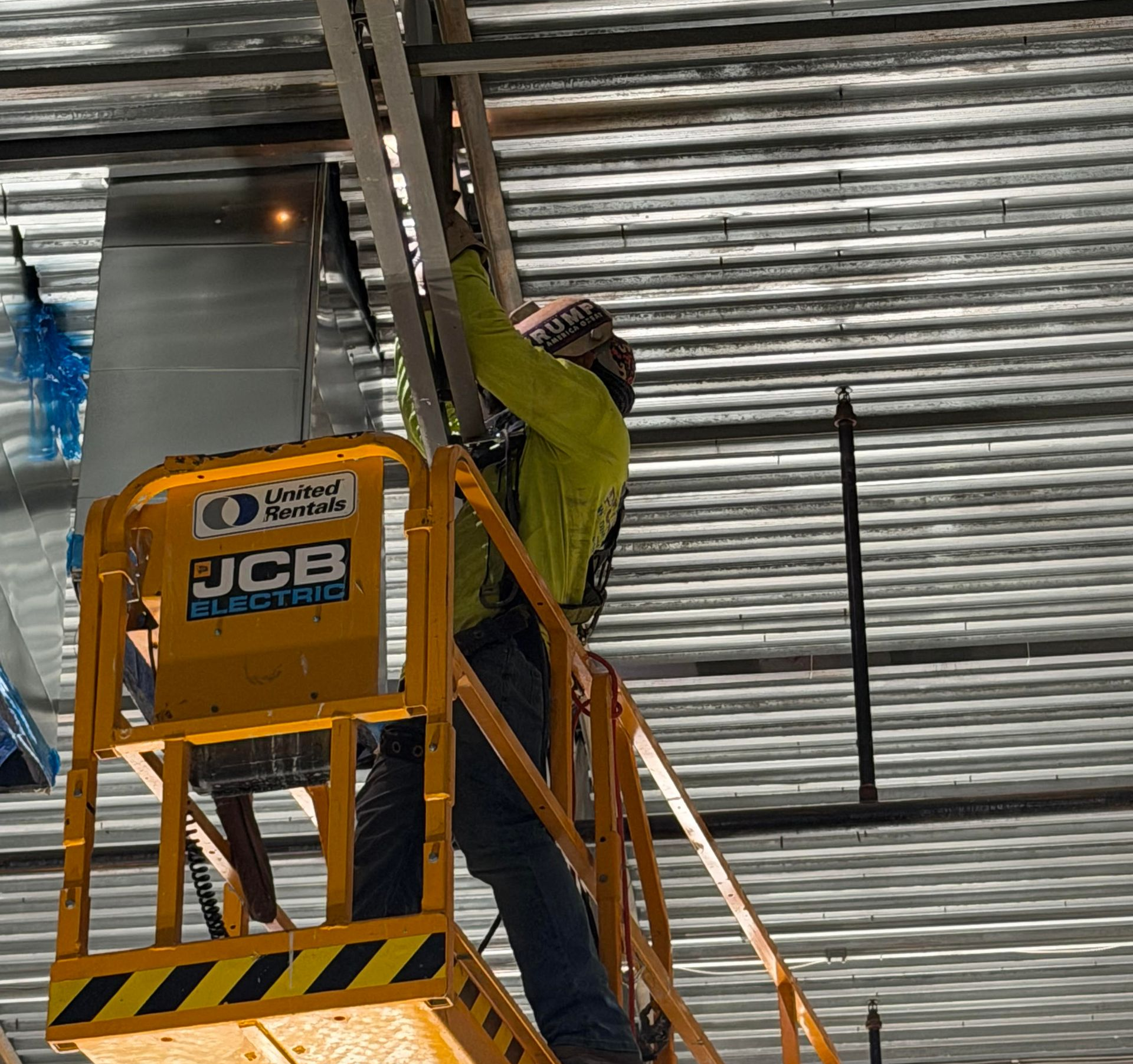 Worker in a yellow lift installs roofing under a corrugated metal roof