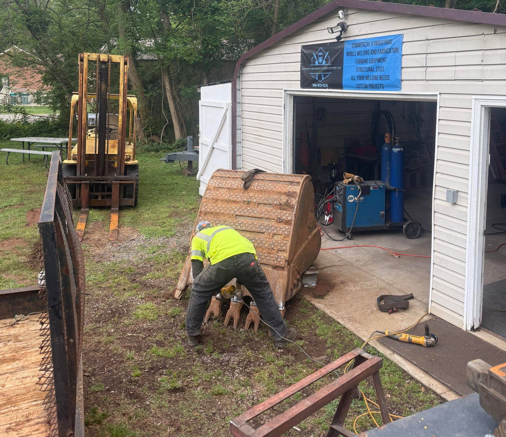 Worker in yellow vest loading a large spool beside a garage and excavator in a muddy yard