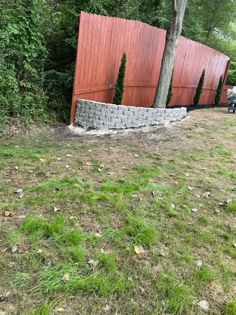 Grassy clearing beside a leaning red fence and low stone wall by dense trees