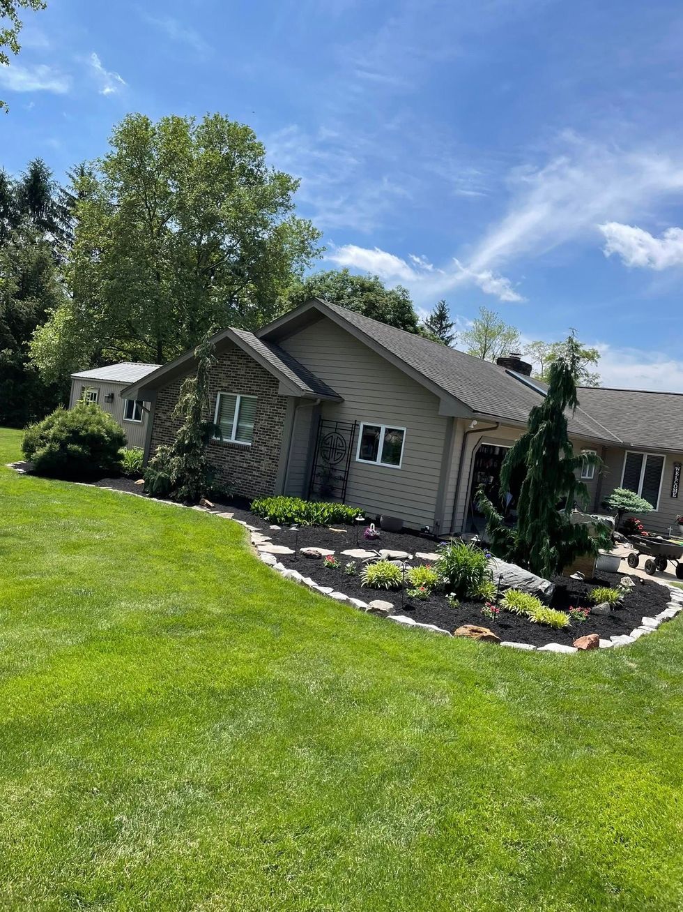 Stone house with gray siding, flower beds, and a lush green lawn under a bright blue sky