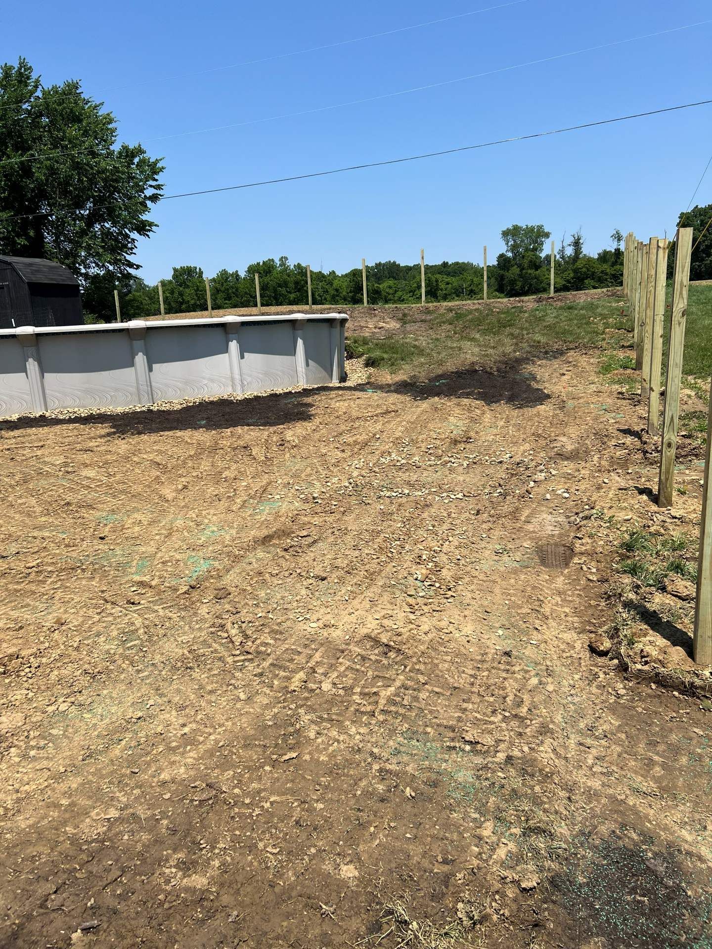Dry dirt lot with a white fence, wooden posts, and a grassy slope under a clear blue sky