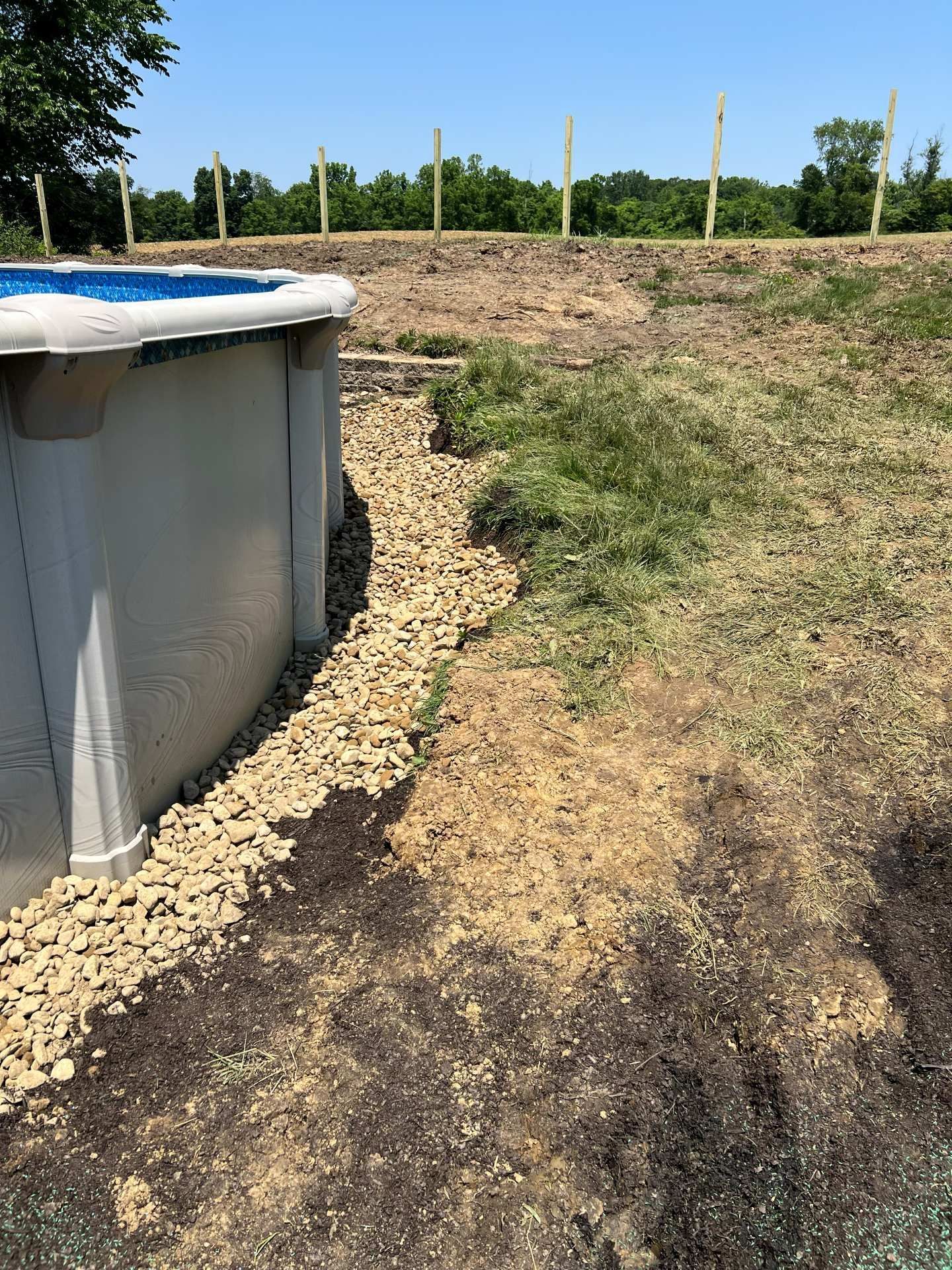 Above-ground pool beside a rocky dirt yard and grassy field under sunny skies