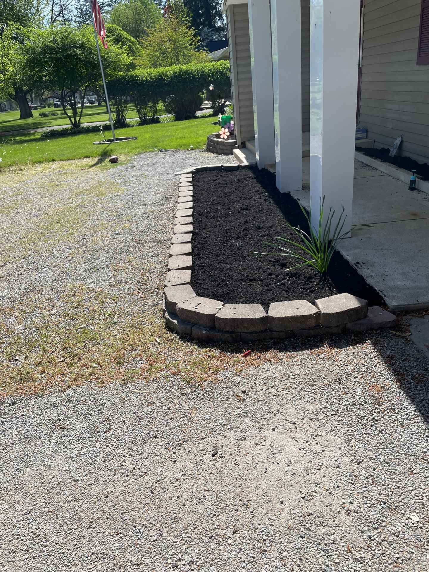 Mulched planting bed with stone border beside white house columns, next to a gravel driveway.