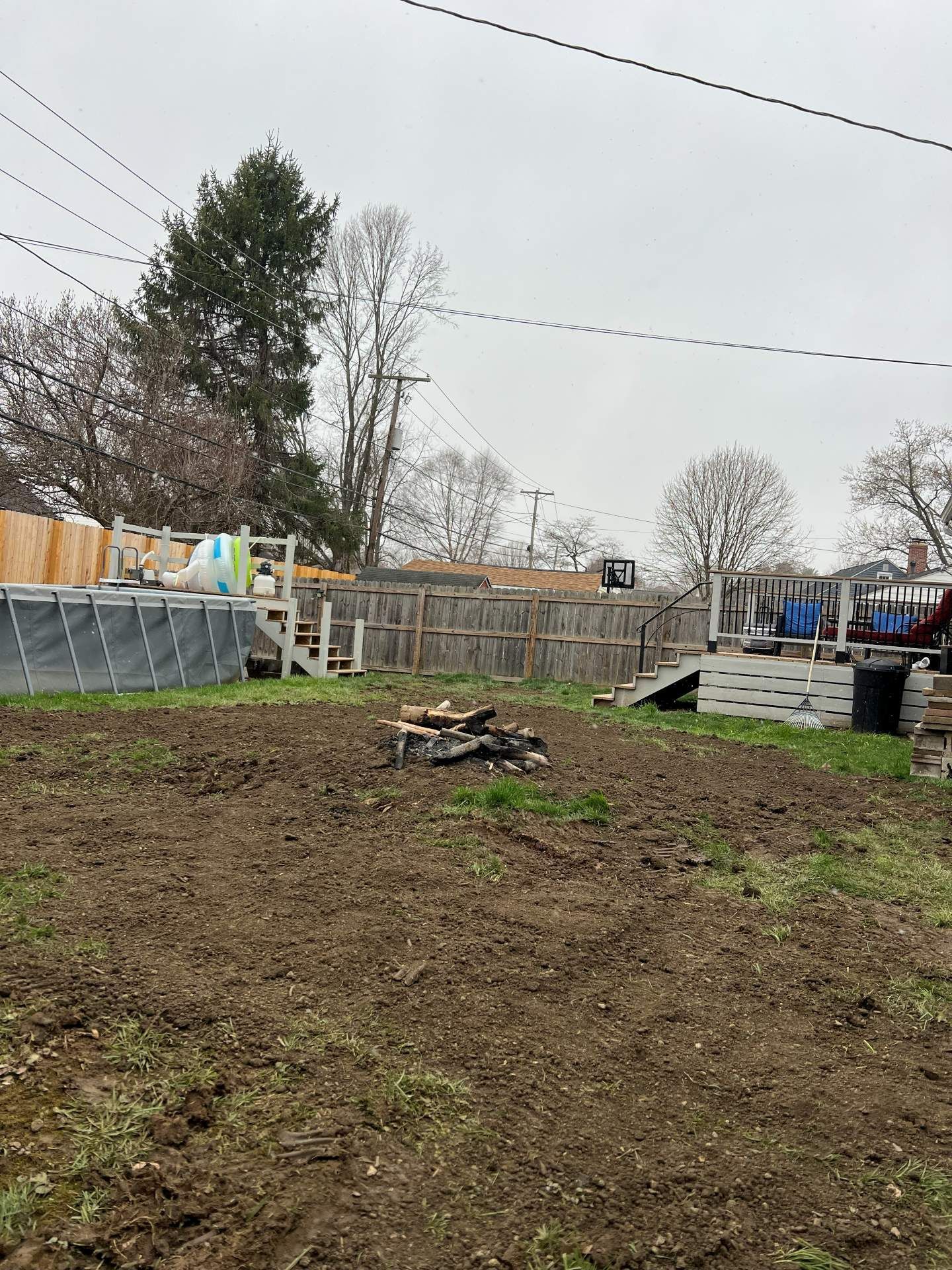 Backyard with muddy ground, fire pit, wooden ramp, fence, trees, and string lights under an overcast sky