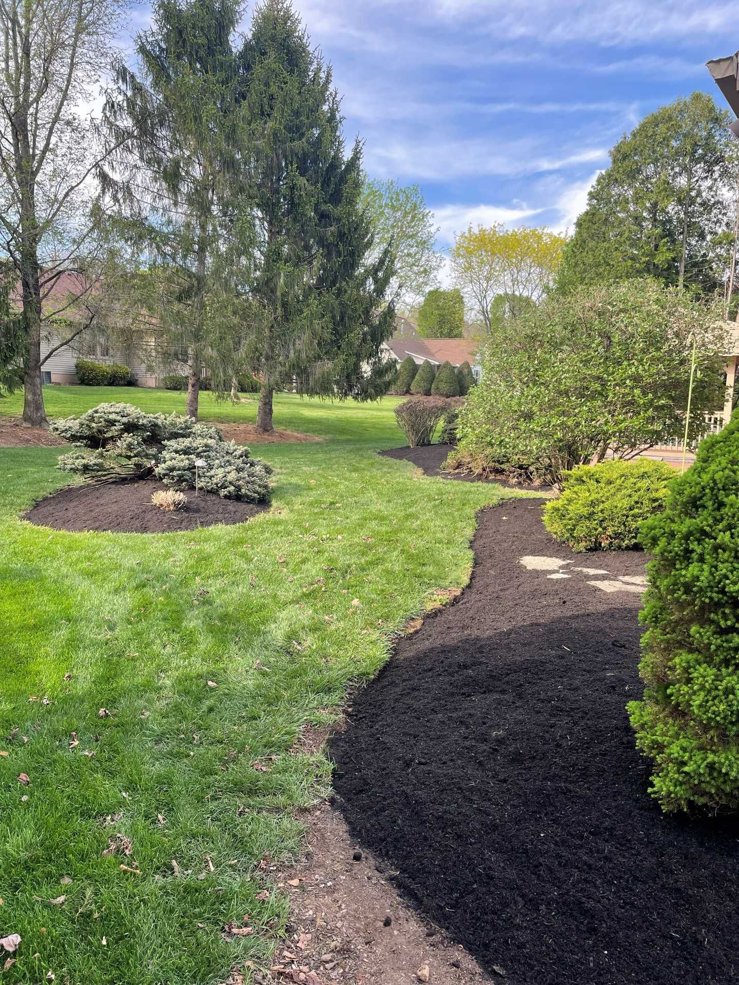 Landscaped garden with green lawn, mulched flower beds, shrubs, and trees under a partly cloudy sky