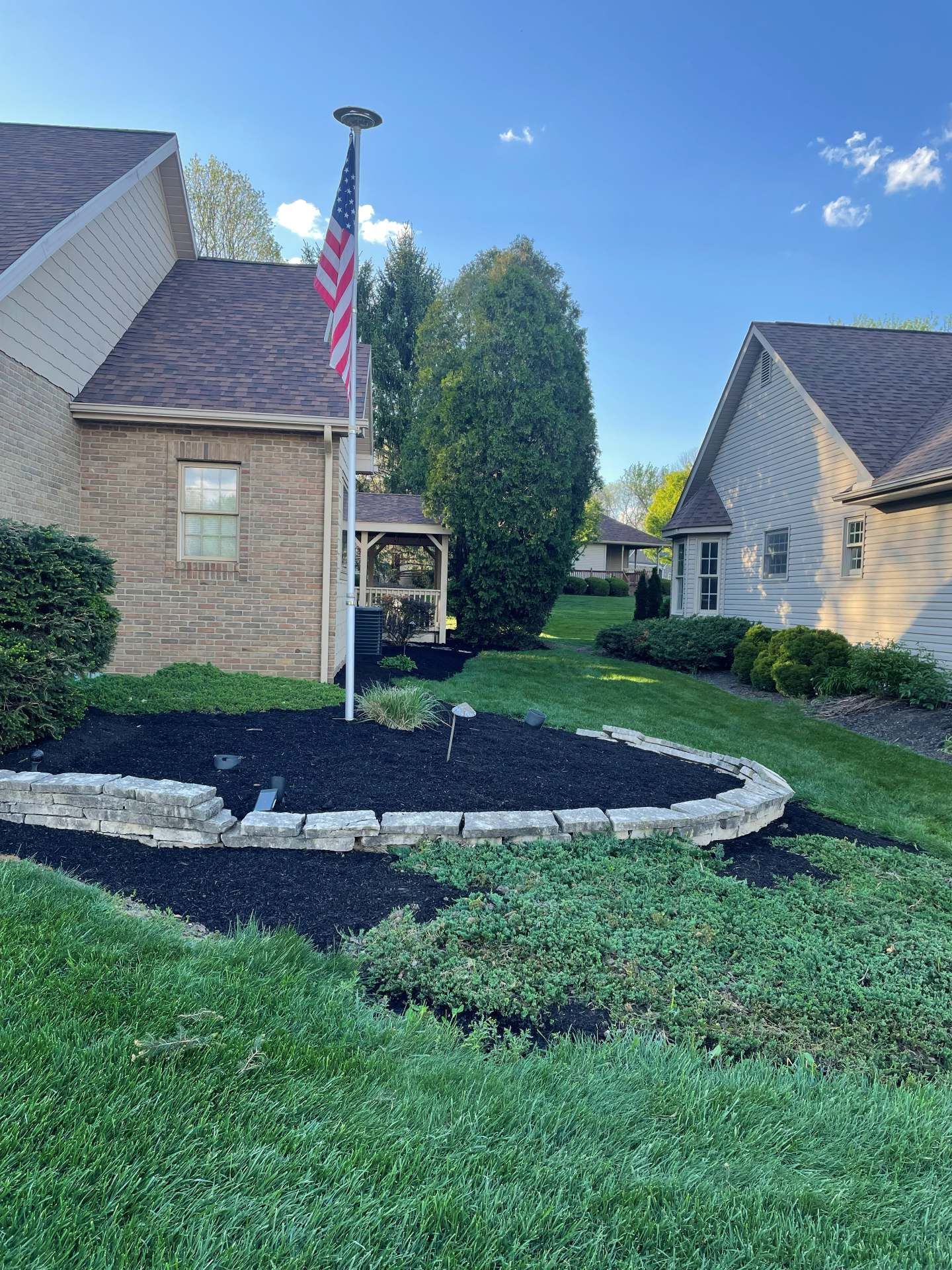 Front yard with brick houses, curved stone border, flagpole, and landscaped mulch beds under a blue sky