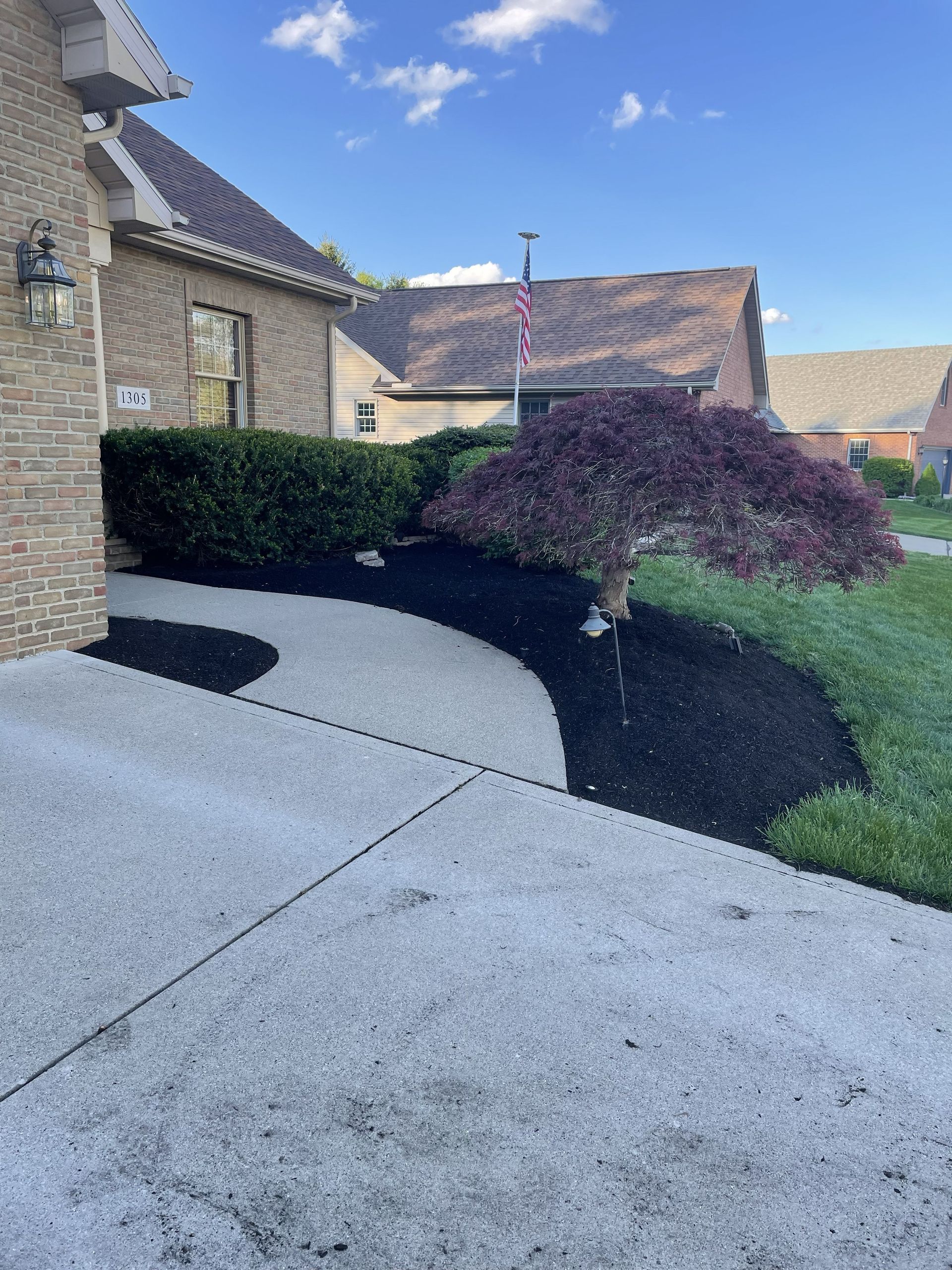 Curved concrete driveway beside stone houses, trimmed hedges, and a burgundy shrub on a sunny day