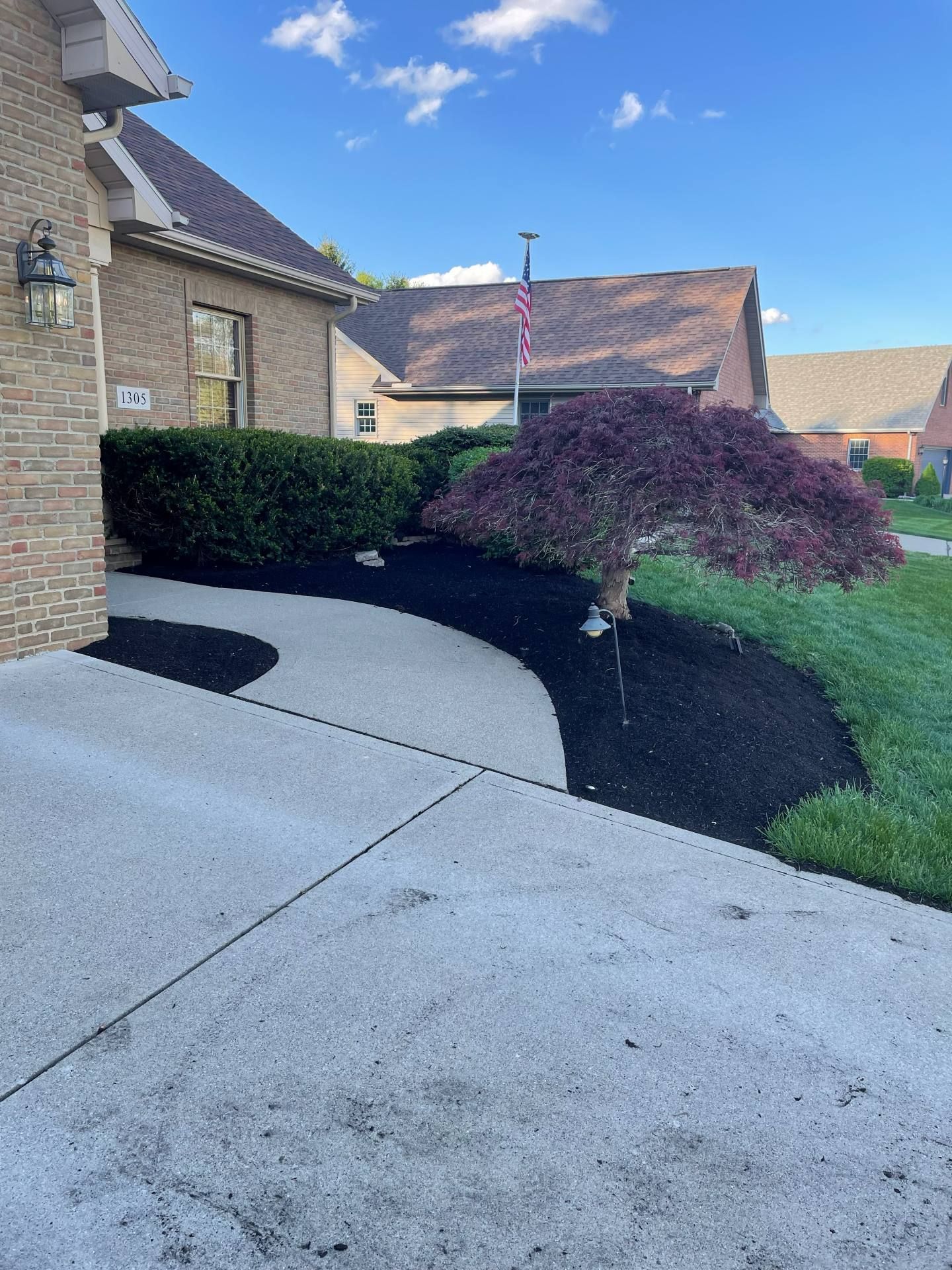 Curved concrete driveway beside a brick house, bordered by shrubs and a red-leaf tree under a blue sky