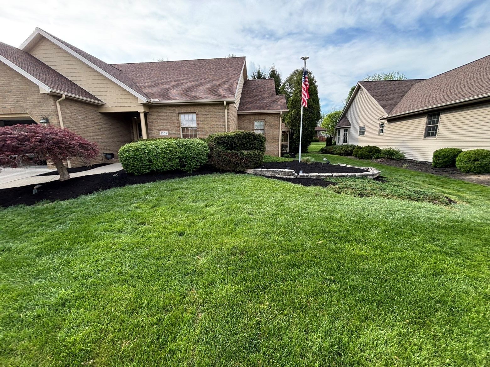 Suburban front yard with green lawn, trimmed shrubs, and tan houses under a cloudy sky