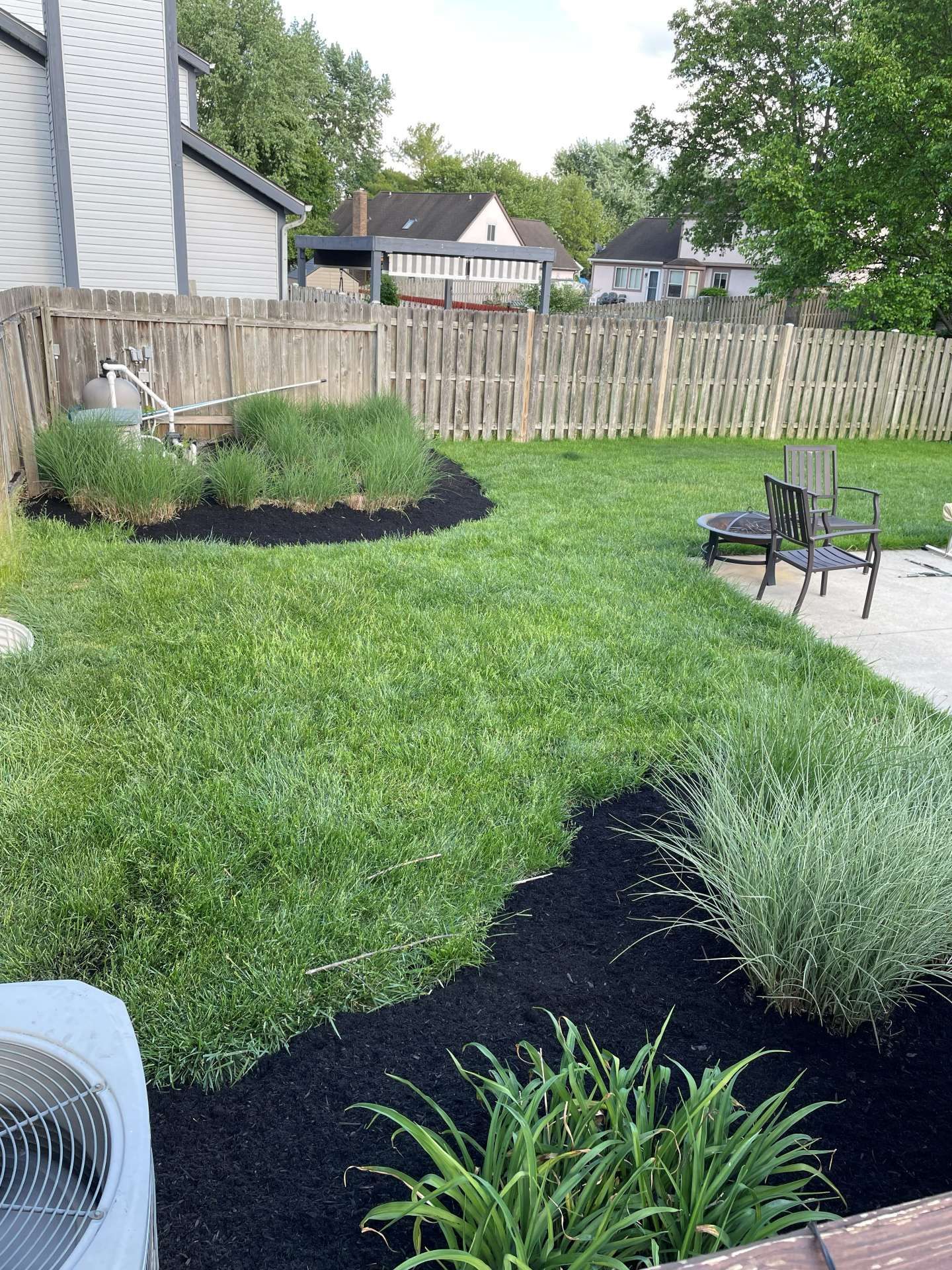 Backyard lawn with black mulch flower beds, a patio chair, and a wooden fence.