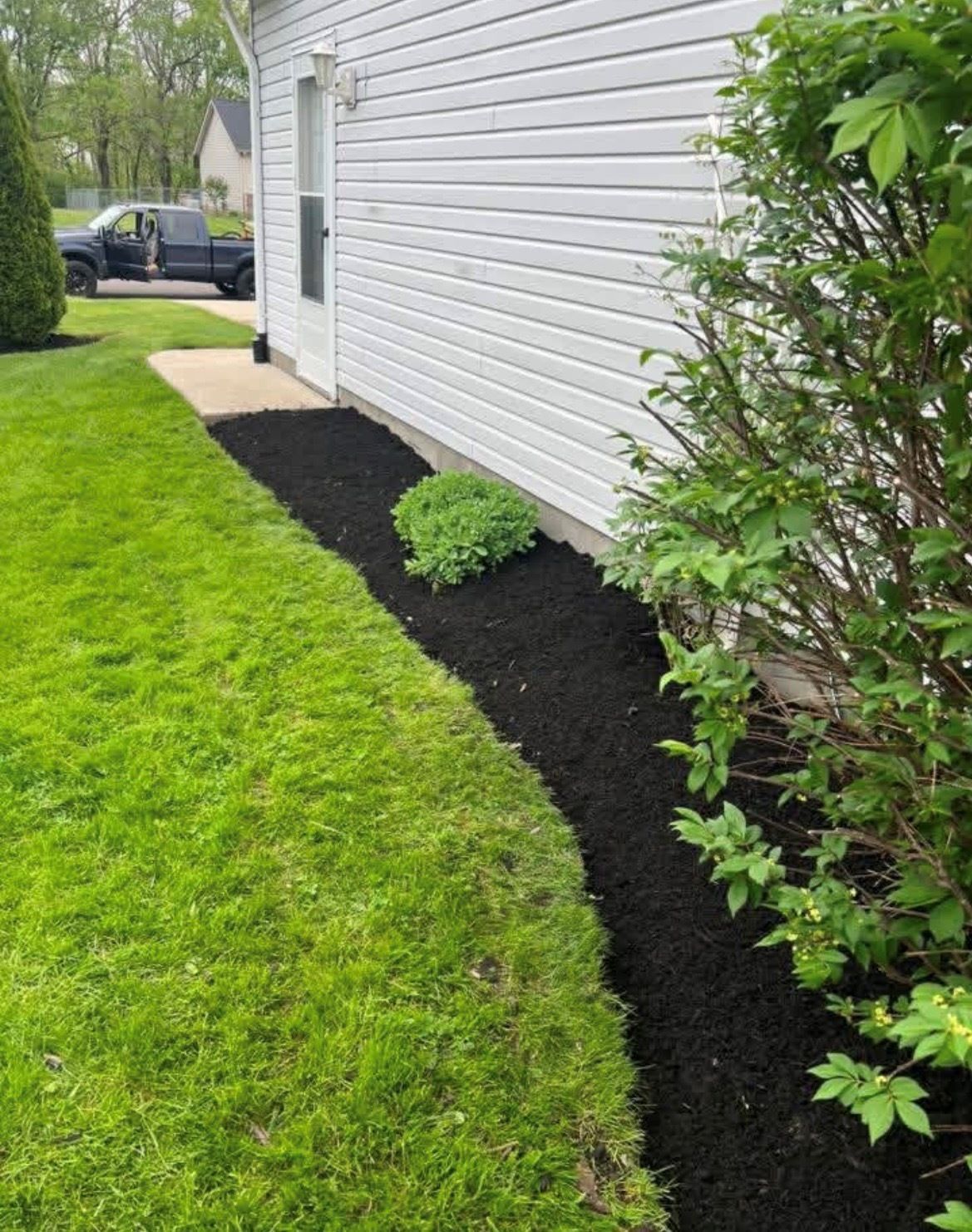 Side yard with fresh black mulch border along a white house and green lawn, with shrubs and a parked car nearby