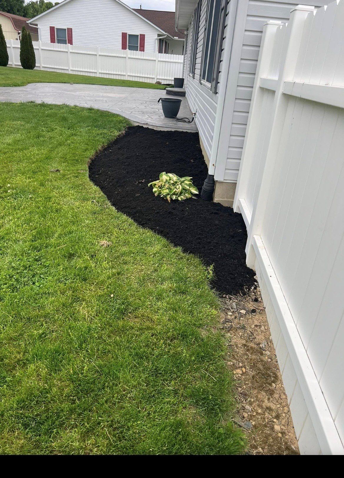 Black mulch bed along house foundation beside white vinyl fence, with grass and a small plant.