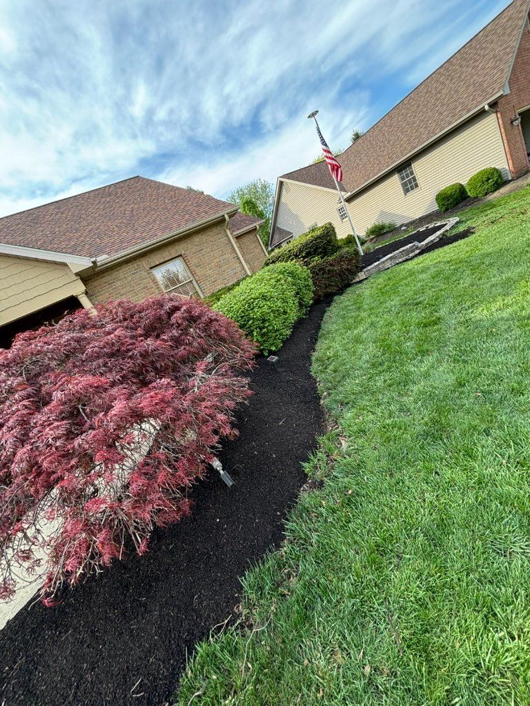Suburban front yard with green lawn, dark mulch beds, pink shrubs, and beige houses under a blue sky