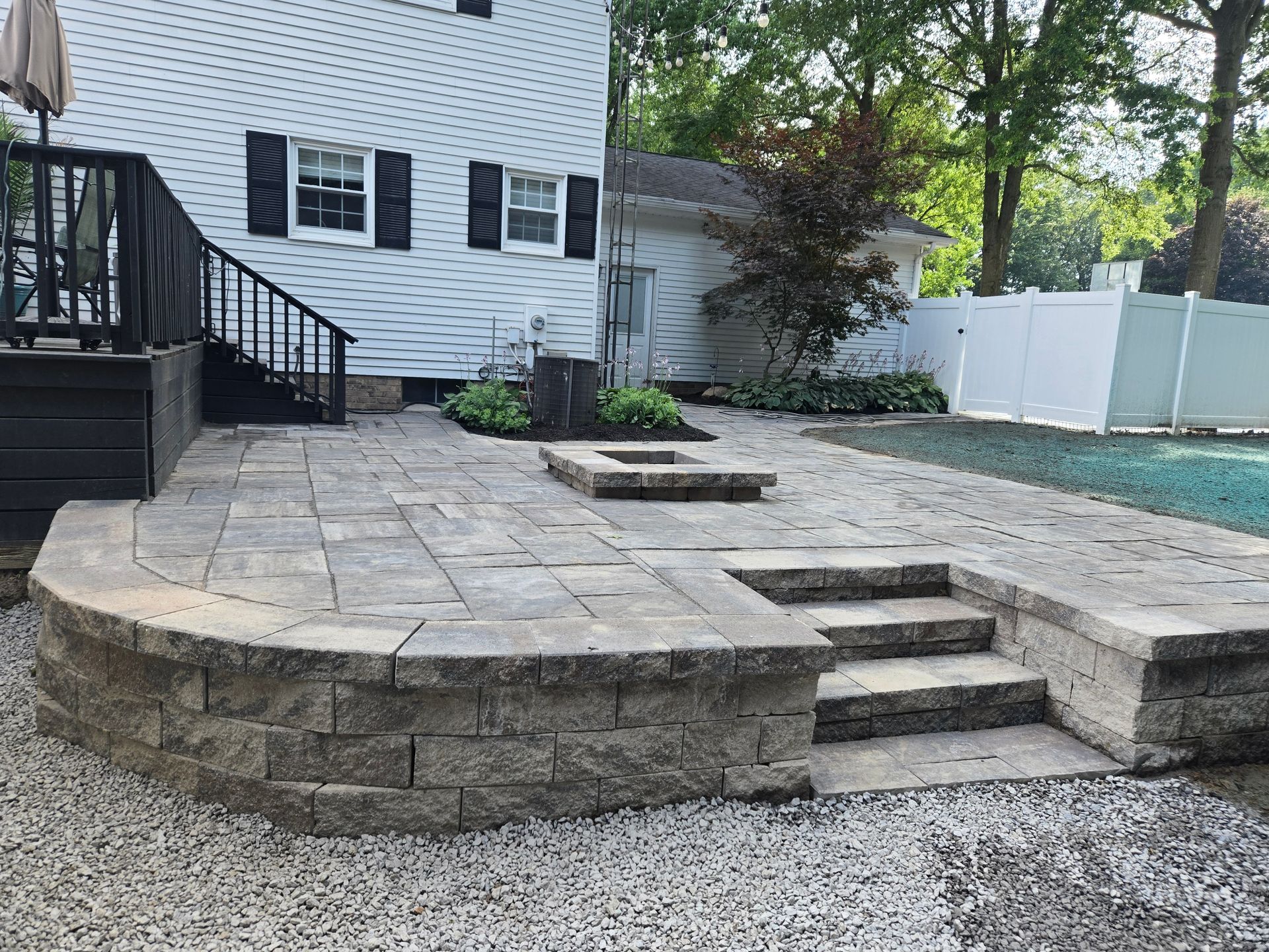 Backyard patio with stone pavers, steps, and a retaining wall. A house and trees are in the background.