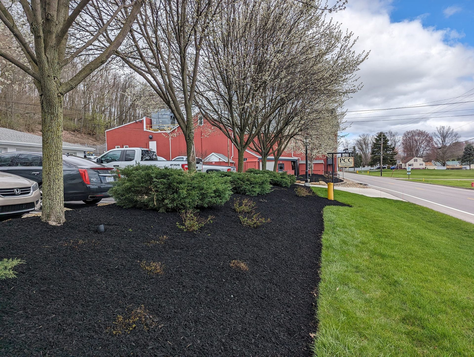 A patch of fresh black mulch and small green shrubs in front of a red building and a road under a partly cloudy sky.