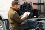 Man in a jacket and cap inspecting a truck, writing on a clipboard.