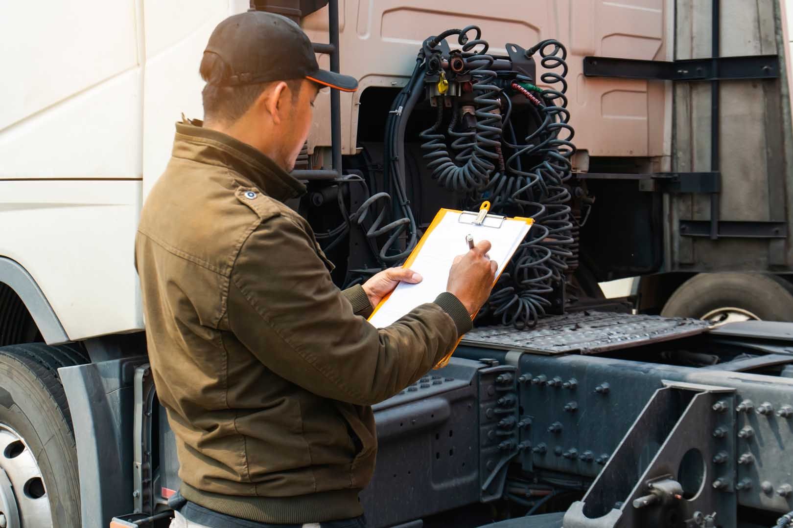Man in a jacket and cap inspecting a truck, writing on a clipboard.