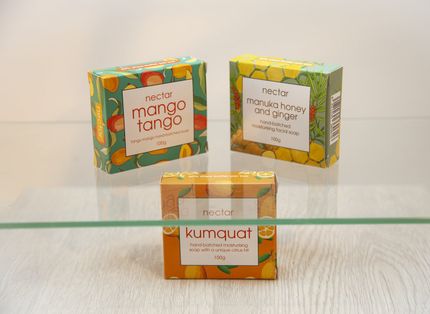 Three colorful soap boxes on a glass shelf: Mango Tango, Manuka Honey & Ginger, and Kumquat.