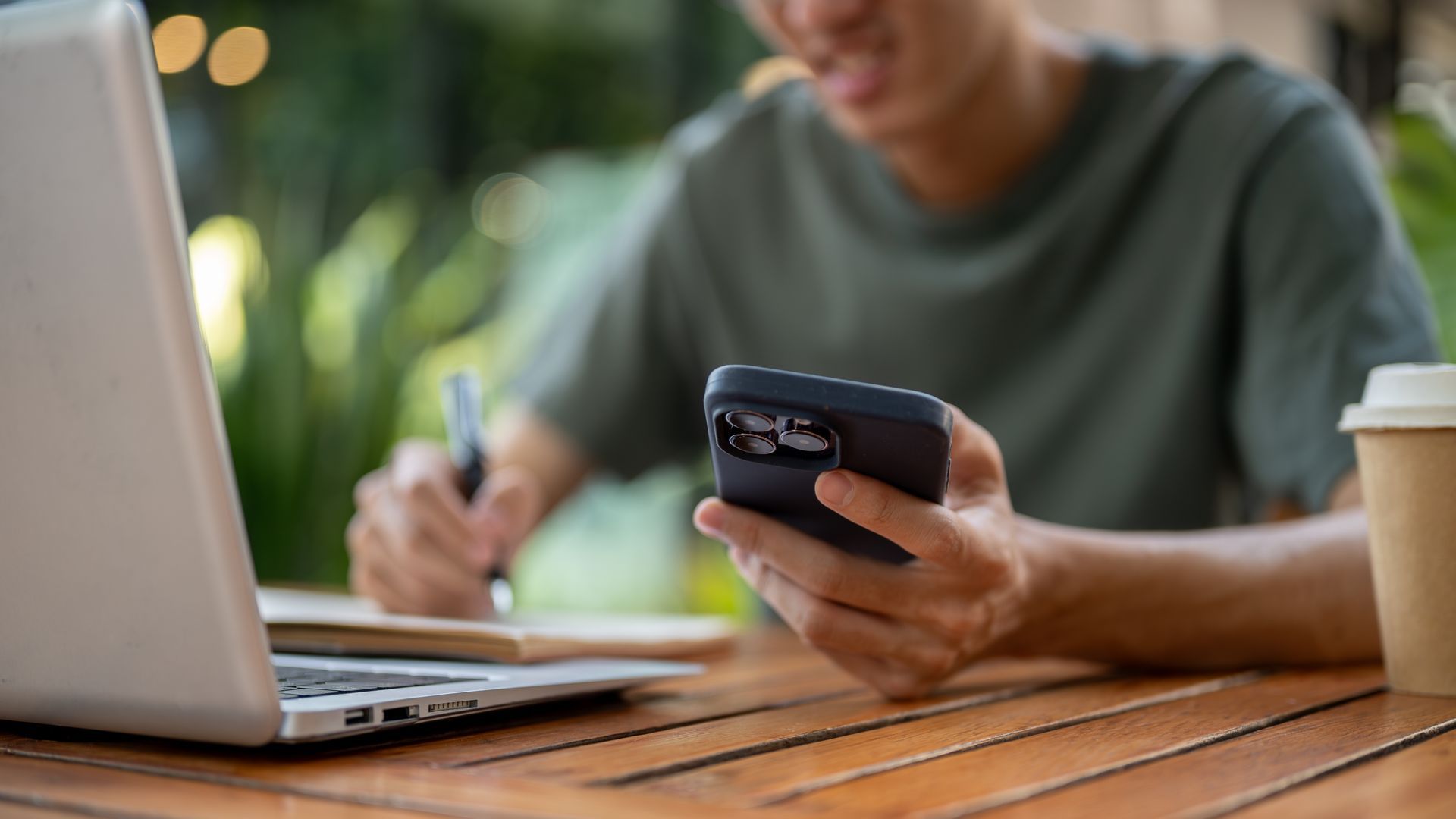 Person using a smartphone at a wooden table beside an open laptop, writing in the background.