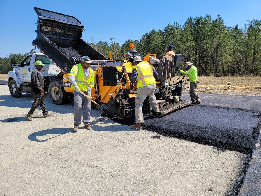 Asphalt paving crew at work; dump truck, paving machine, workers in safety vests, blue sky.