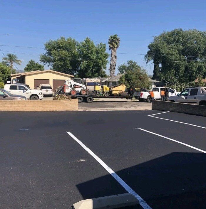 Asphalt parking lot with construction vehicles and trailers in front of buildings on a sunny day.
