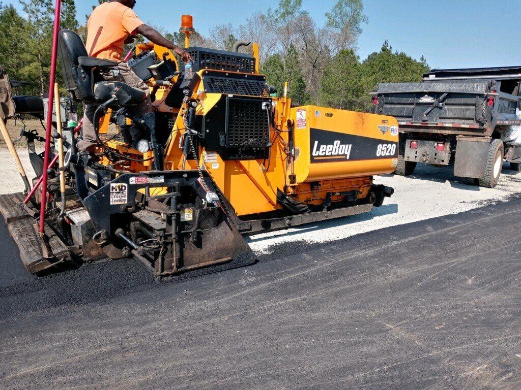Road paving machine laying asphalt; worker operating controls; truck nearby.