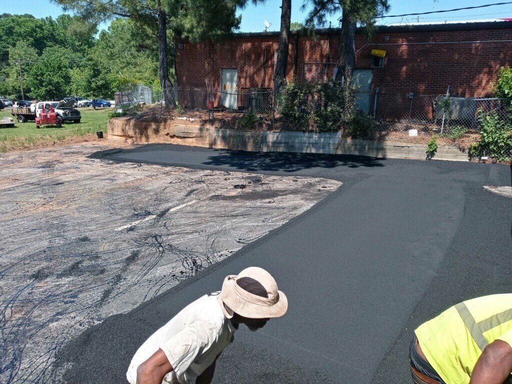 Workers paving a parking lot with fresh asphalt. One wears a straw hat, another a safety vest, under a bright sky.