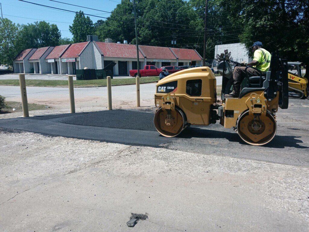 Asphalt roller compacting fresh pavement; worker operates machine in front of low building.