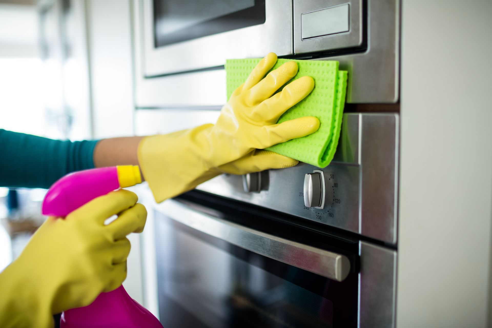 Gloved hands cleaning a stainless steel oven with green cloth and pink spray bottle