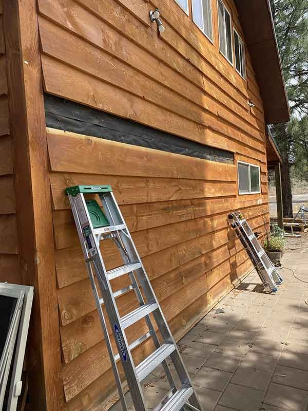 Two ladders leaning against a brown wooden house exterior beside a paved walkway.