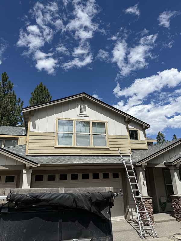 Two-story house under repair with ladder and tarp, against a bright blue sky with clouds.