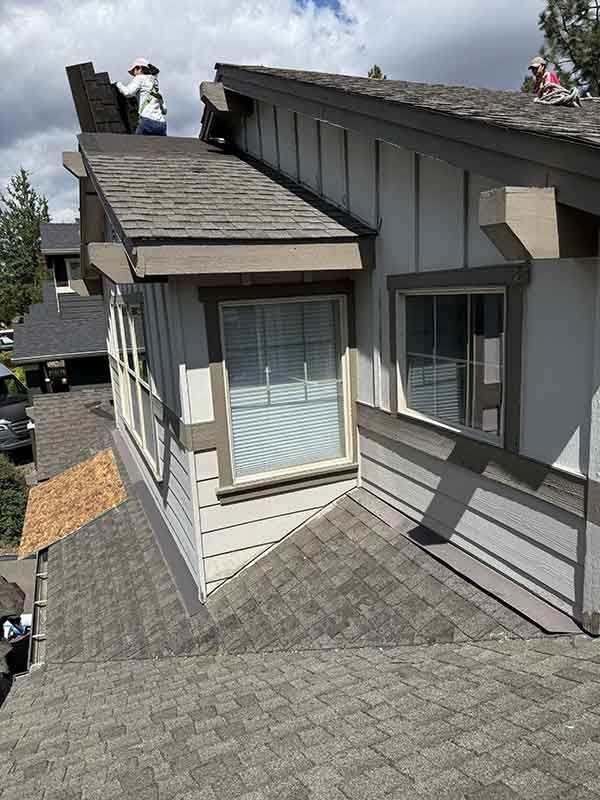 Rooftop shingle repair on a gray suburban house, with a worker near the chimney.